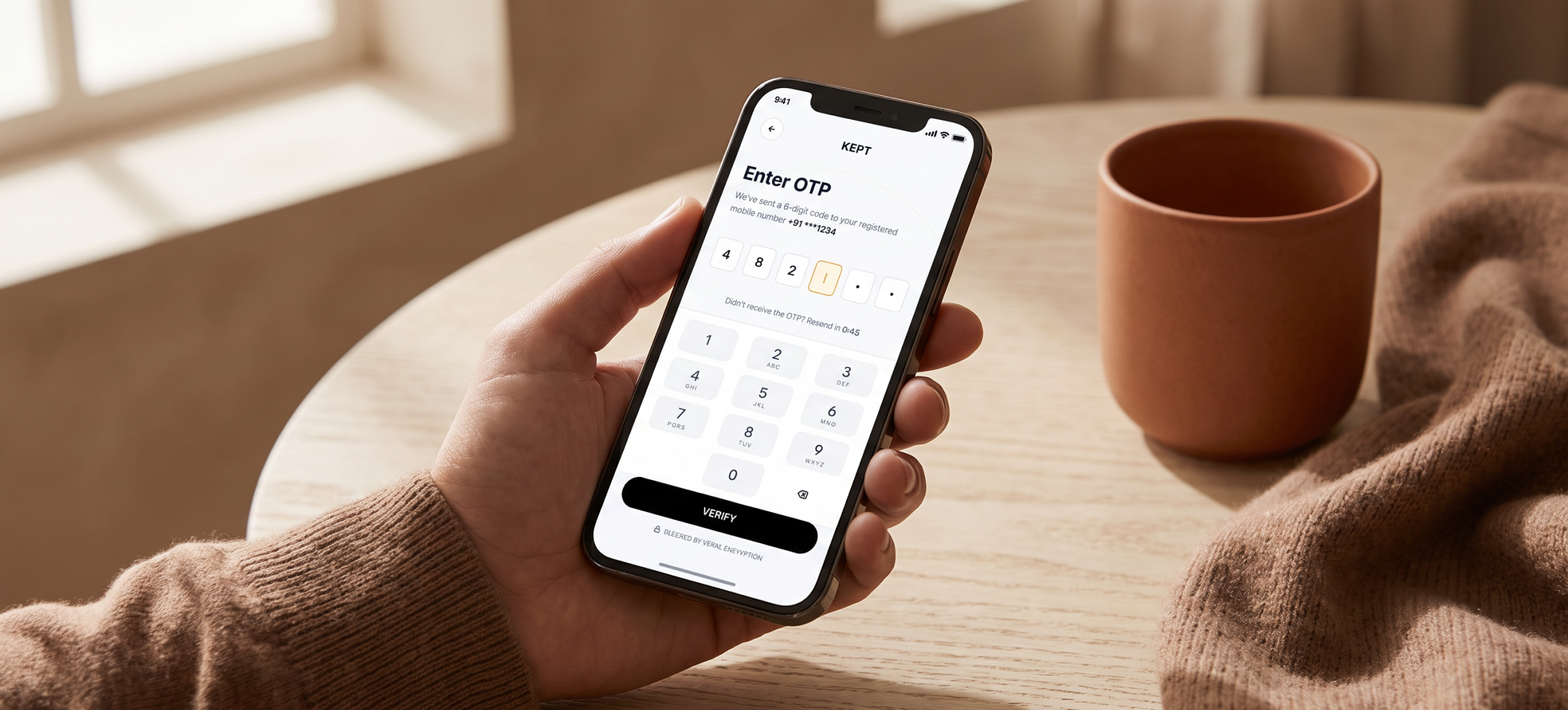 A hand holding a phone showing the Kept OTP screen at a kitchen table
